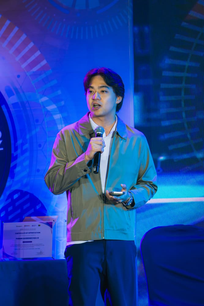 A speaker in a jacket holding a phone and speaking into a microphone on stage, with a certificate of appreciation displayed beside him.