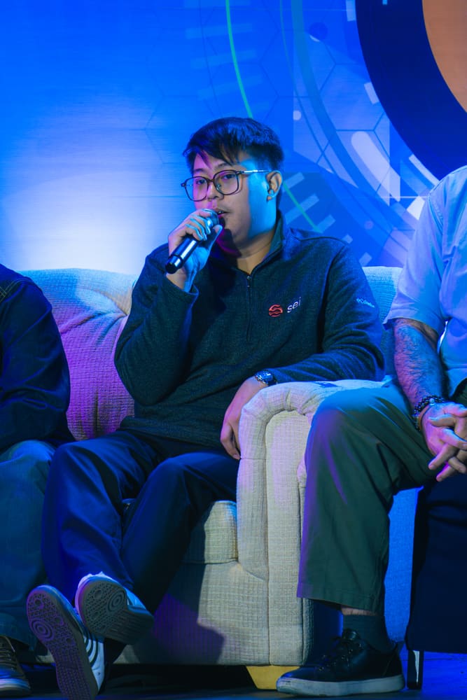A panelist wearing glasses and a fleece jacket, speaking into a microphone from a couch during a panel discussion.