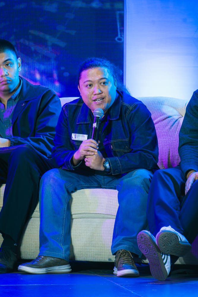 A panelist in a denim jacket wearing a name badge, speaking into a microphone on a couch during a panel discussion.