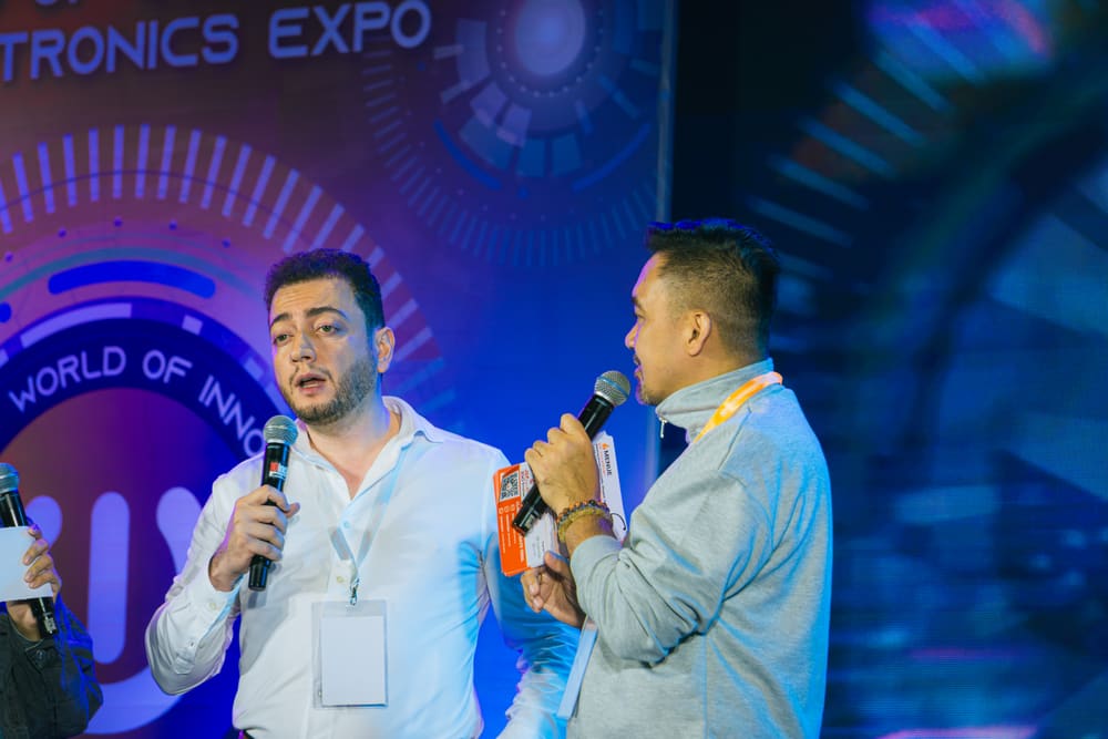 Two speakers standing close together sharing microphones on an expo stage backdrop reading "World of Innovation".