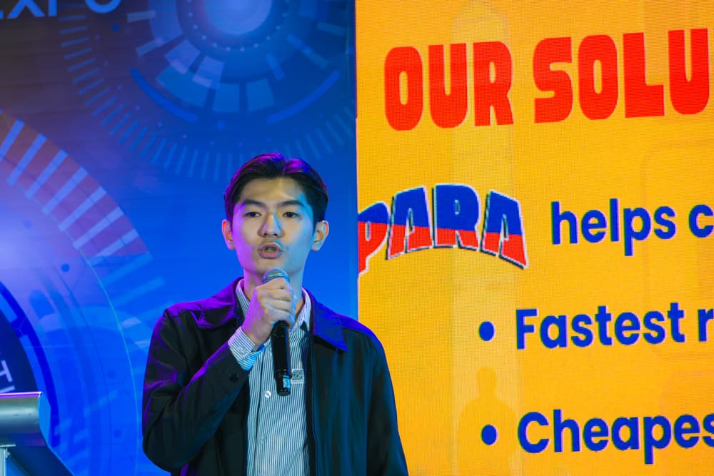 A speaker presenting beside a bright yellow slide about a company's solutions during an expo talk.