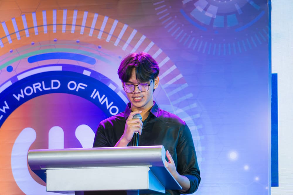A speaker at a podium smiling while speaking into a microphone under purple stage lighting.