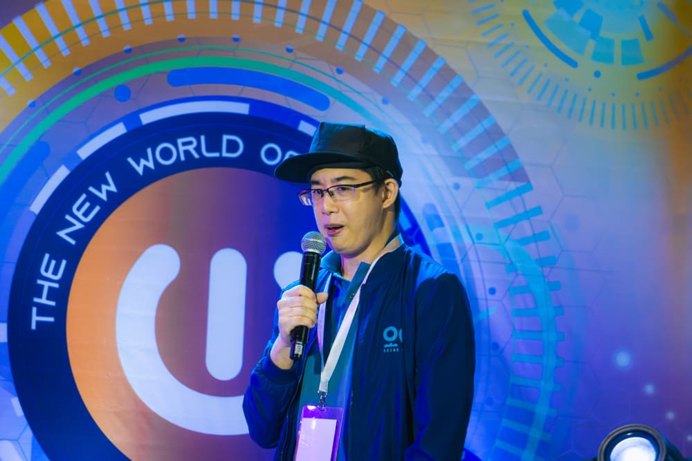 A speaker wearing a cap and a jacket with an ocean-themed logo, speaking into a microphone on an expo stage.